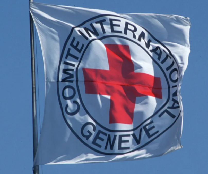 ธง ICRC รูปกากบาทสีแดงบนพื้นหลังสีขาว มีวงกลมล้อมรอบเขียนว่า COMITÉ INTERNATIONAL GENÈVE กำลัง ปลิวไสวในสายลม ด้านหลังเป็นท้องฟ้า